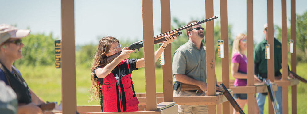 Young woman in a red shooting vest aiming a shotgun at five-stand wooden stands