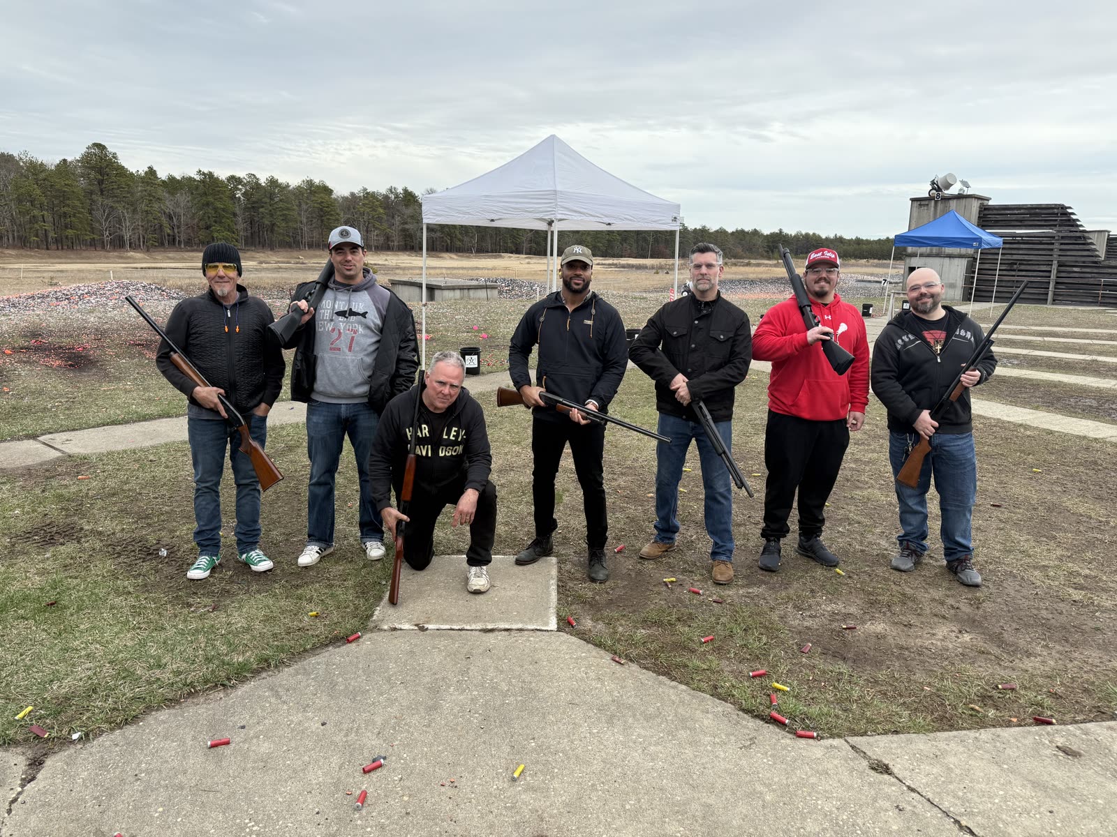 Seven friends on the range with shotguns, one kneeling in a Harley hoodie