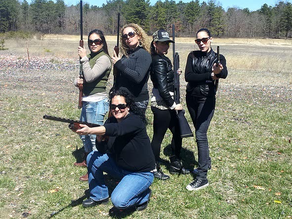 Five women posing with shotguns on a sunny clay field